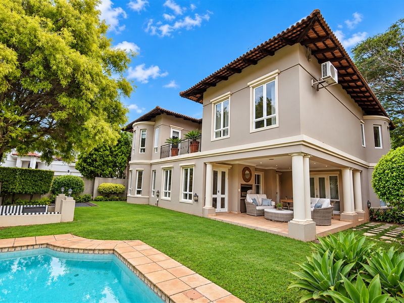 Rear view of a two-story house featuring a covered patio with a built-in braai, a swimming pool, and a landscaped garden.