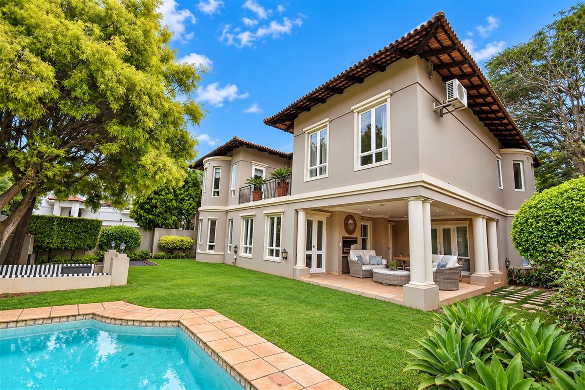 Rear view of a two-story house featuring a covered patio with a built-in braai, a swimming pool, and a landscaped garden.