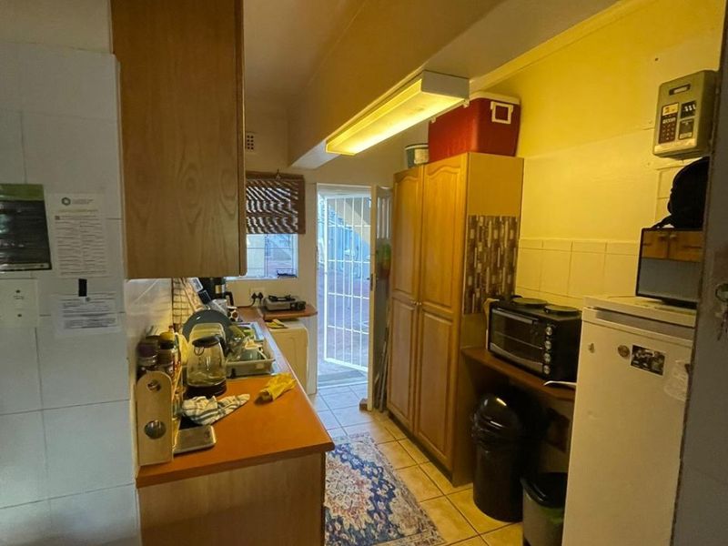 A compact kitchen featuring wooden cabinetry, laminate countertops, and tiled flooring, leading to an exterior door. The space would benefit from modern updates.