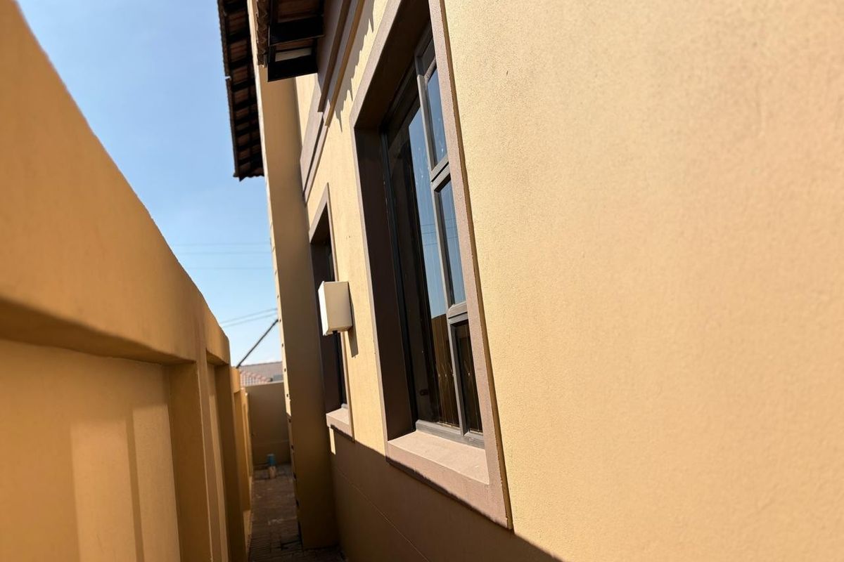A side passage view showing the exterior wall of the house, paved walkway, boundary wall, and aluminium window frames.