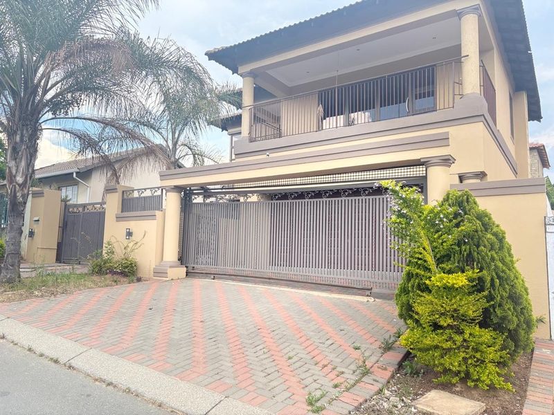 Front elevation of a double-story house featuring a paved driveway, secure sliding gate, and an upper-level balcony with metal railings.