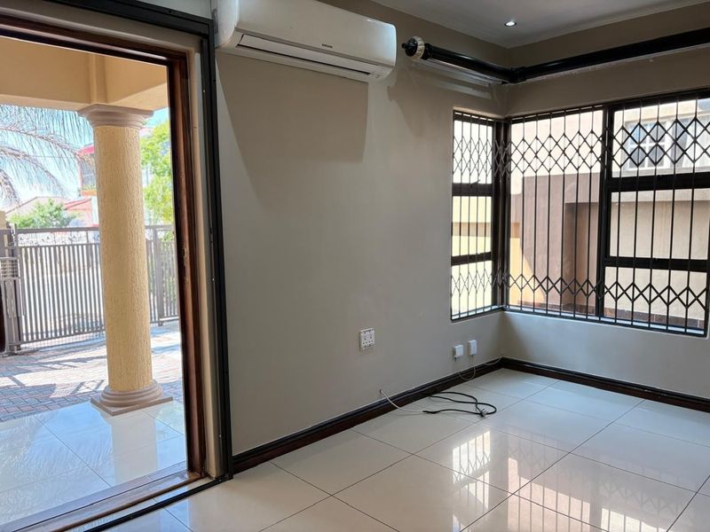 A bright, empty living area featuring tiled floors, a wall-mounted air conditioning unit, and security gates on the windows, with direct access to a patio.