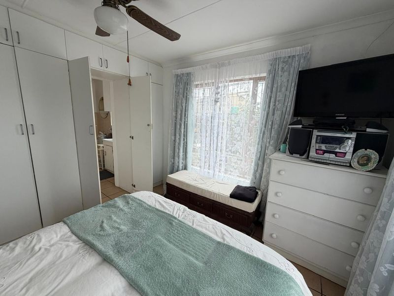 A functional bedroom featuring built-in cupboards, a ceiling fan, and security bars on the window, with direct access to an en-suite bathroom.
