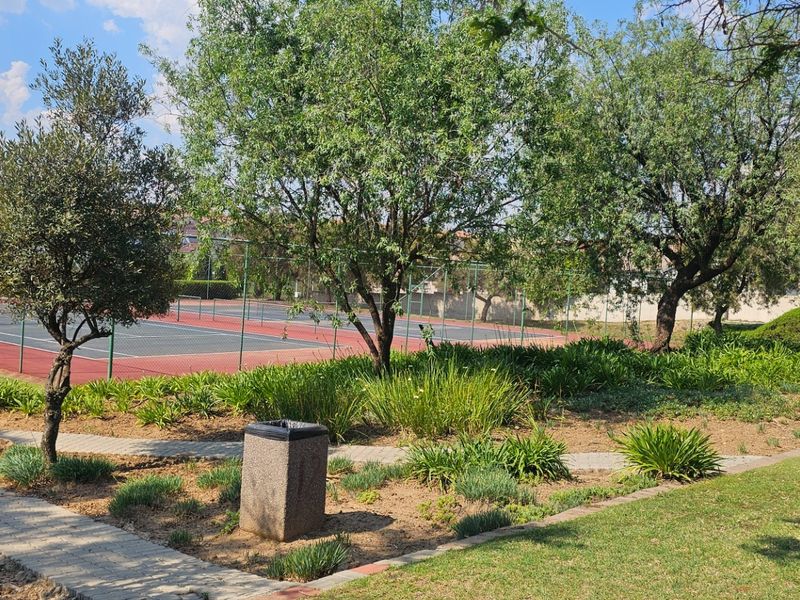 A well-maintained communal tennis court facility surrounded by landscaped gardens and paved pathways within the residential estate.