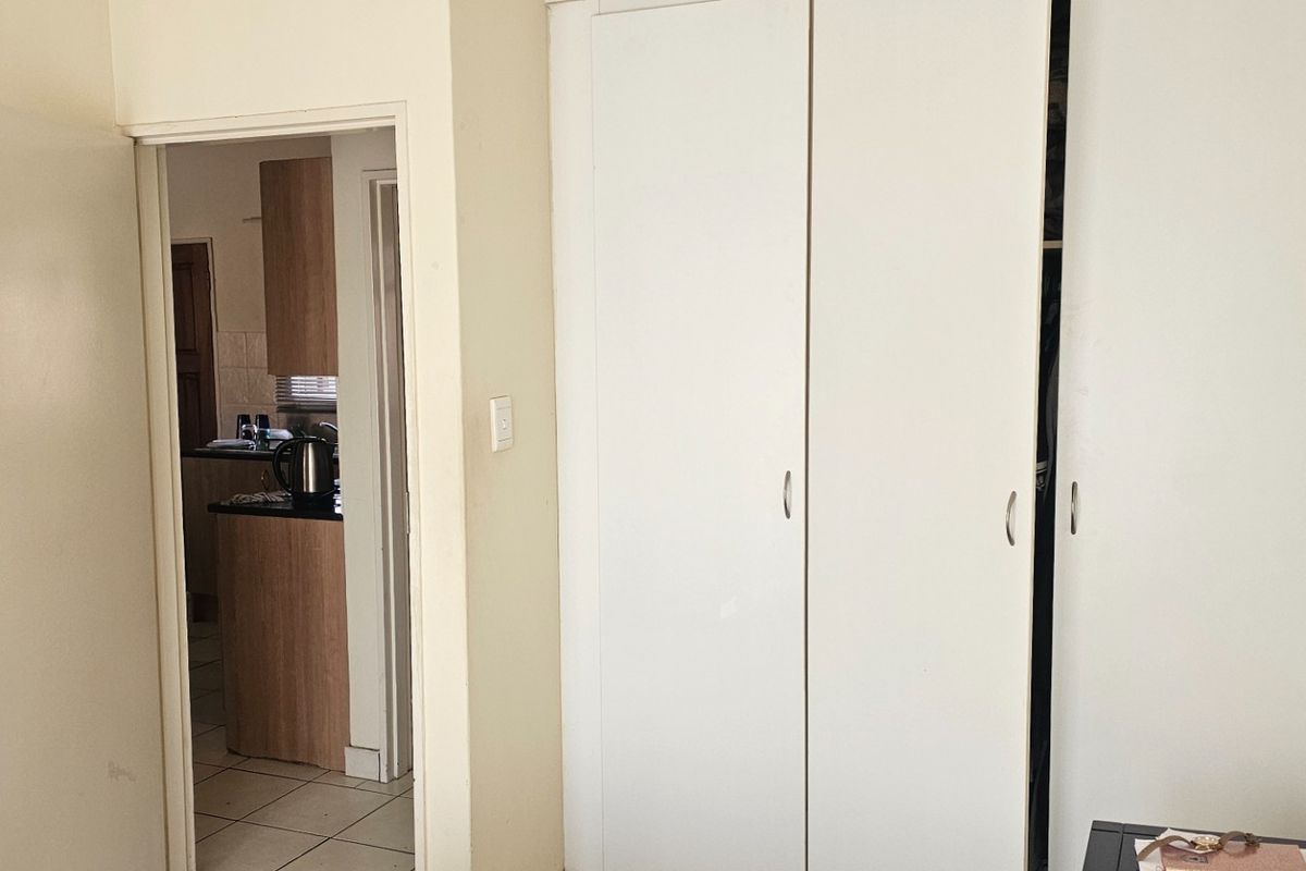 A bedroom featuring built-in cupboards and tiled flooring, with a view through the doorway into the adjacent kitchen area.
