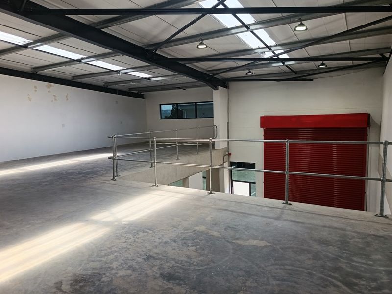 Spacious industrial mezzanine level featuring exposed steel trusses, skylights, concrete flooring, and a secure red roller shutter door, ideal for storage or workspace.