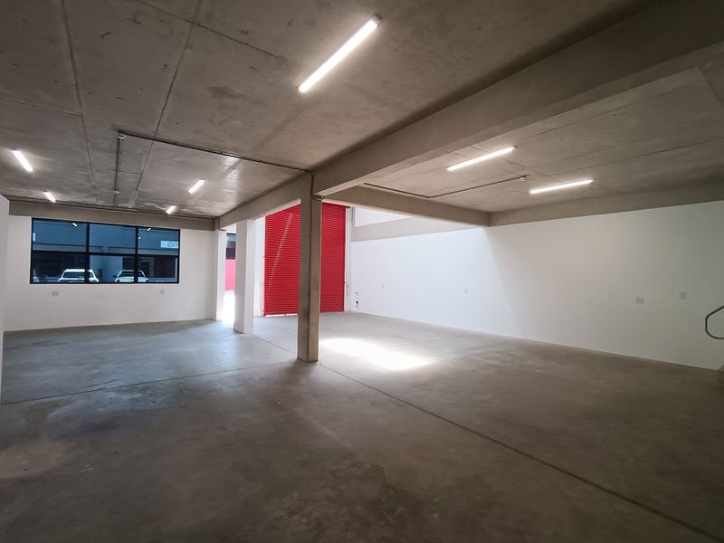 Spacious industrial utility area featuring concrete flooring, exposed concrete ceilings, bright fluorescent lighting, and a large red roller shutter door.