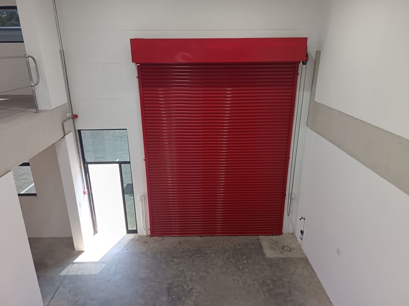 A clean, modern industrial warehouse space featuring a prominent red roller shutter door, concrete flooring, and an elevated mezzanine level with metal safety railing.