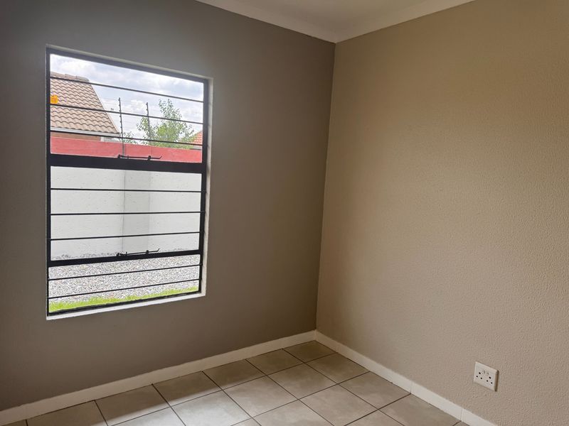 A clean, empty bedroom featuring neutral-toned walls, tiled flooring, and a window fitted with security bars.