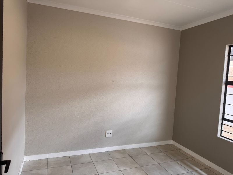 A clean, empty bedroom featuring neutral painted walls, tiled flooring, and a window fitted with security bars.