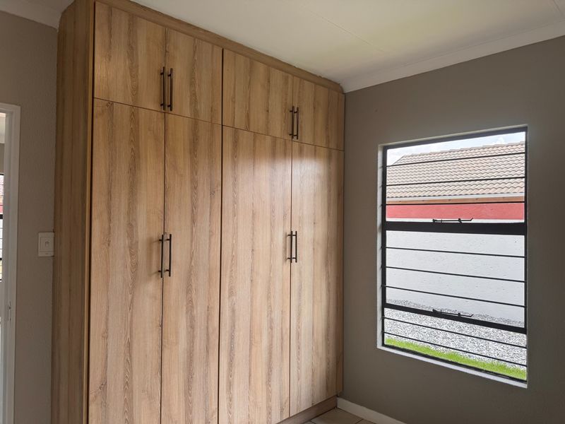 A clean bedroom featuring modern wood-finish built-in cupboards and a window with security bars, offering a neutral and well-maintained space.