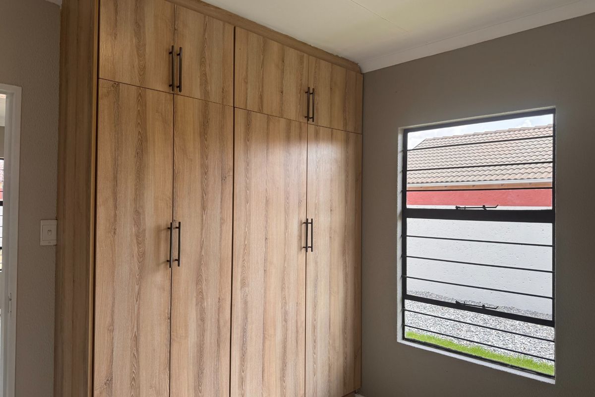 A clean bedroom featuring modern wood-finish built-in cupboards and a window with security bars, offering a neutral and well-maintained space.