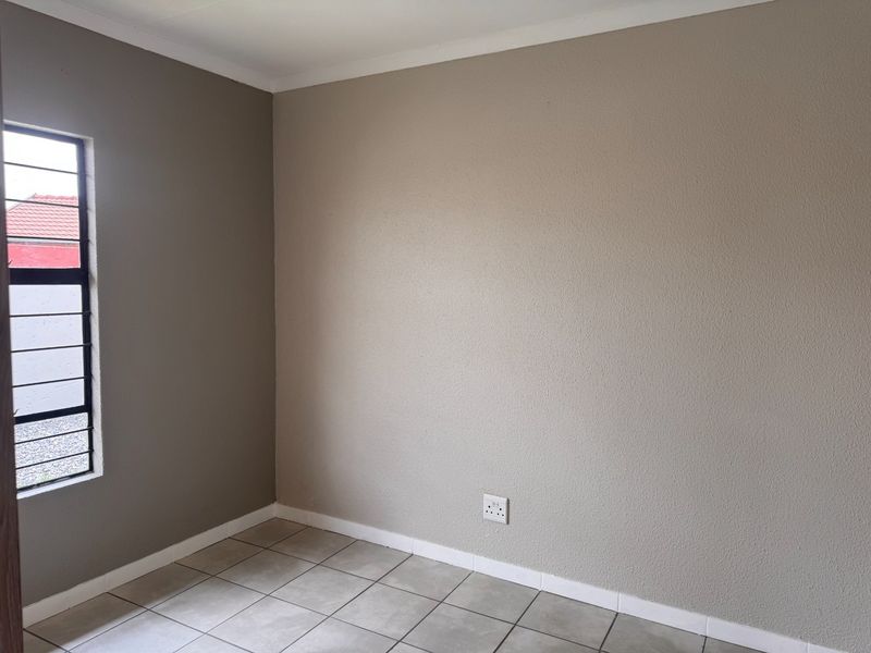 A clean, empty bedroom featuring neutral-toned walls, tiled flooring, and a window fitted with security bars.