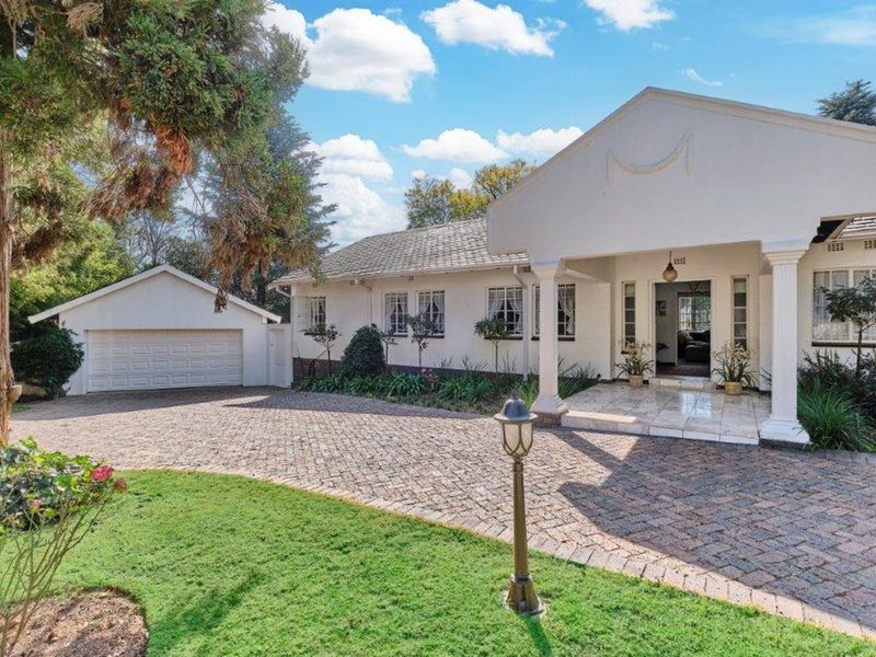 A charming front elevation featuring a paved driveway, detached garage, and a welcoming covered entrance with classical columns and well-maintained garden surroundings.