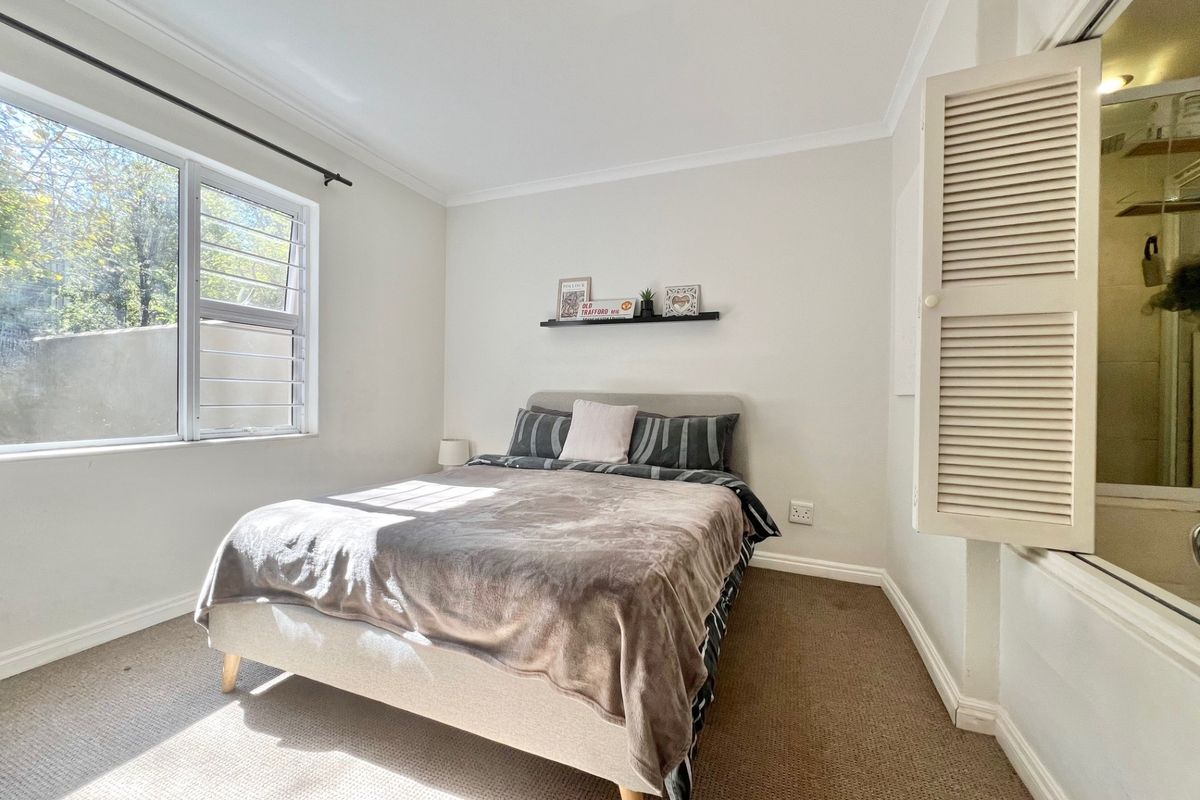 A well-lit bedroom featuring carpeted flooring, a louvered window, and an internal shutter window providing a view into an adjacent space.