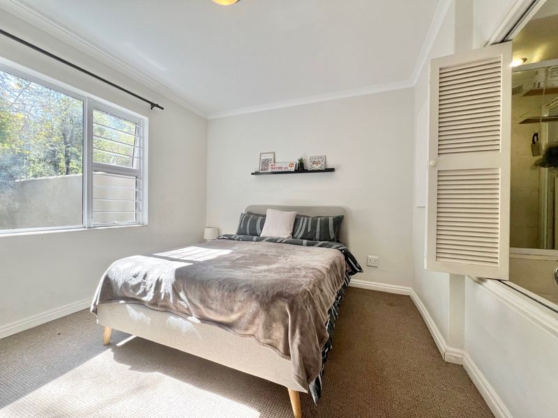 A well-lit bedroom featuring carpeted flooring, a louvered window, and an internal shutter window providing a view into an adjacent space.