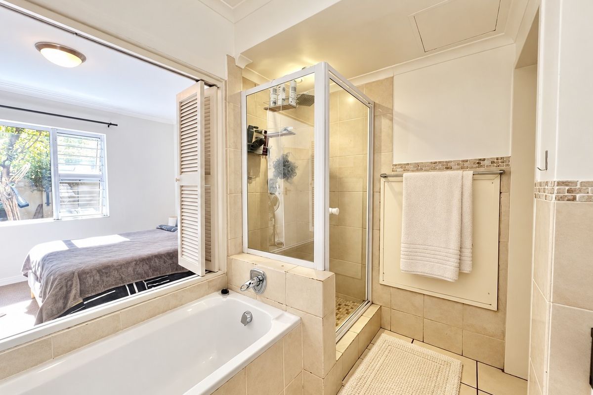 A functional bathroom featuring a built-in bathtub, a separate glass-enclosed shower, and neutral wall and floor tiling with a view into the adjacent bedroom.
