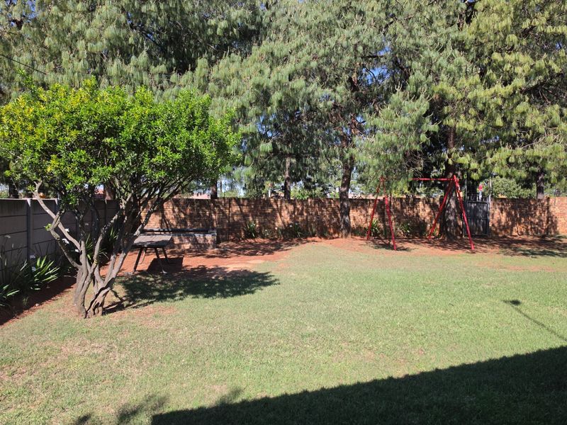 A spacious backyard garden featuring a well-maintained lawn, mature trees, a brick boundary wall, and a built-in braai area.