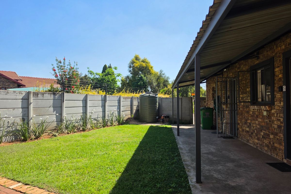 A rear exterior view featuring a covered concrete patio, a brick facade, a lawn area, and a precast concrete boundary wall with electric fencing.