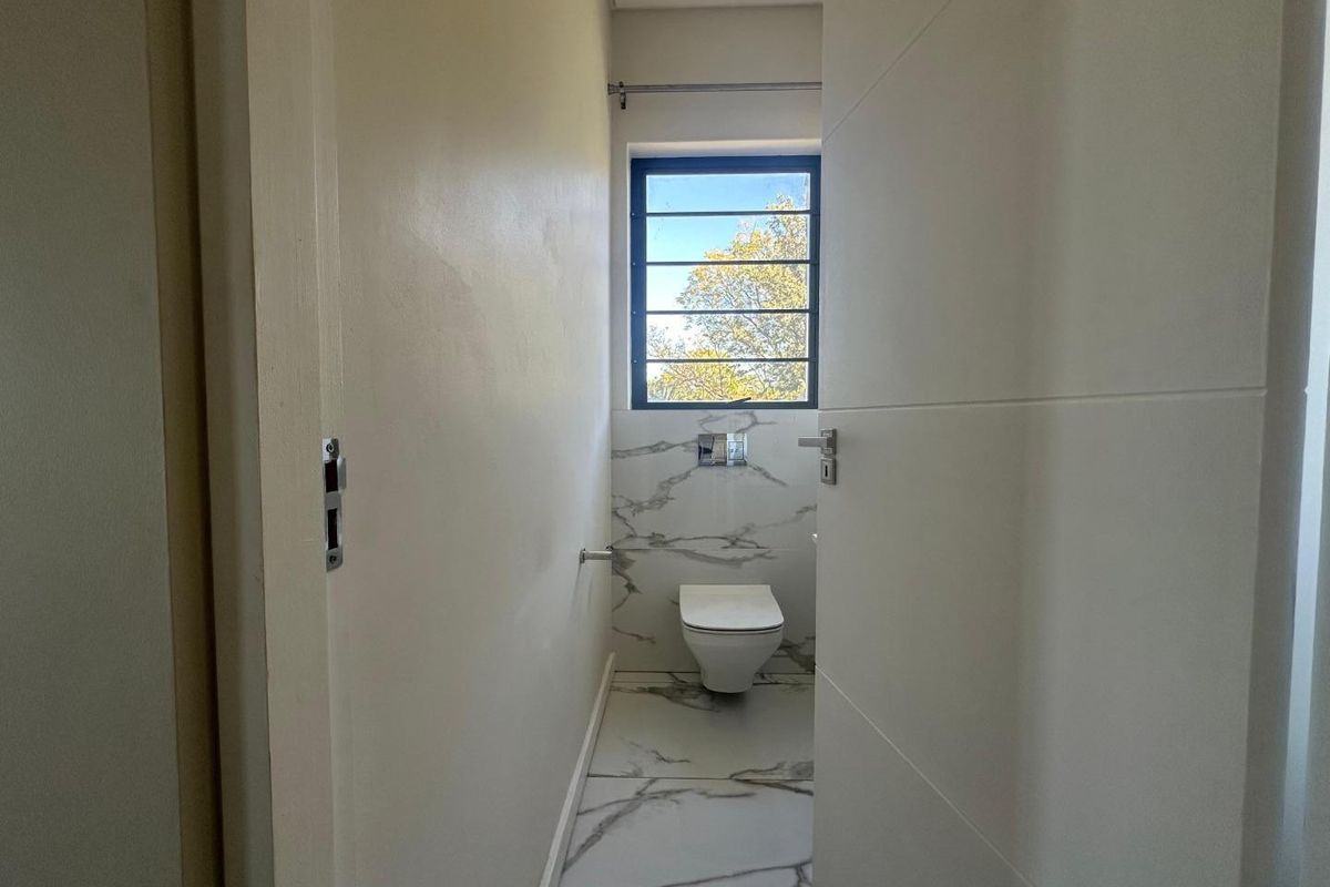 A modern, minimalist guest bathroom featuring marble-look floor and wall tiles, a wall-mounted toilet, and a window with security bars.