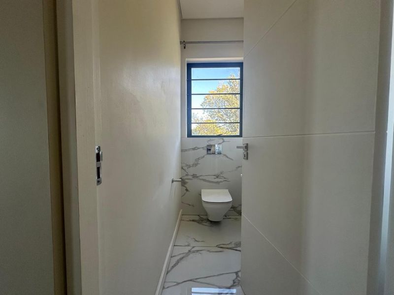 A modern, minimalist guest bathroom featuring marble-look floor and wall tiles, a wall-mounted toilet, and a window with security bars.