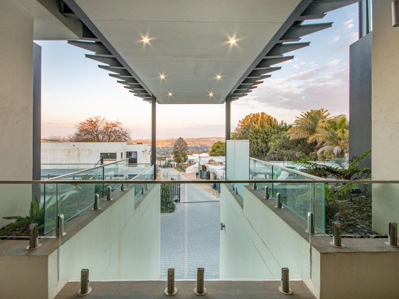 Modern covered balcony featuring a sleek glass balustrade and recessed lighting, offering an elevated view over the property entrance and surrounding landscape.