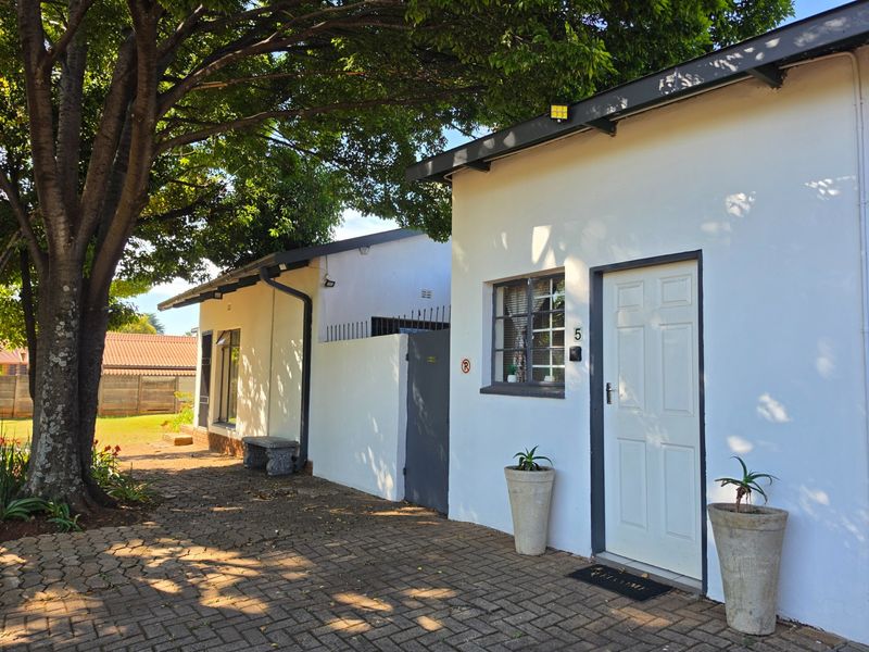 Exterior view of a guest house unit featuring a paved driveway, white painted walls, and secure entrance door with steel window frames.