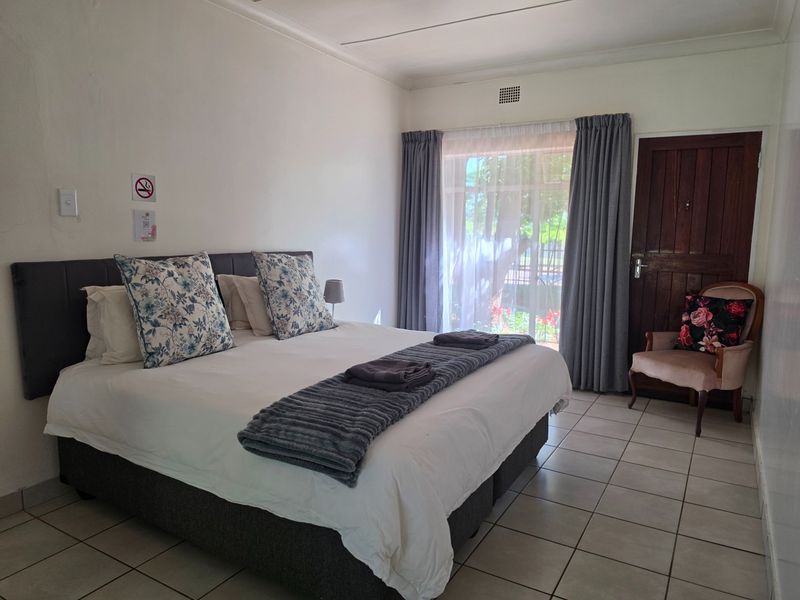 A functional bedroom featuring tiled flooring, a wooden entrance door, and large windows with curtains, providing a simple and clean guest accommodation space.
