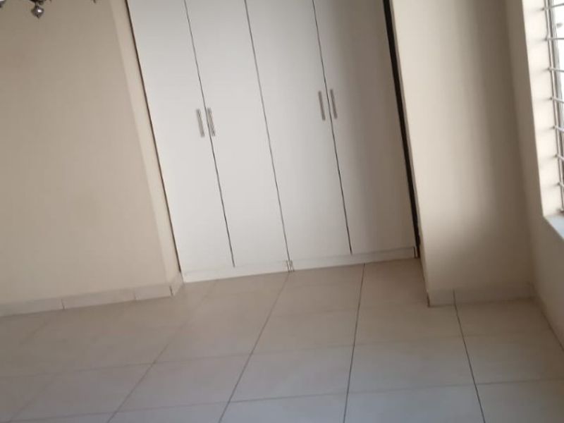A bright, empty bedroom featuring neutral tiled flooring, built-in white cupboards, and security bars on the window.
