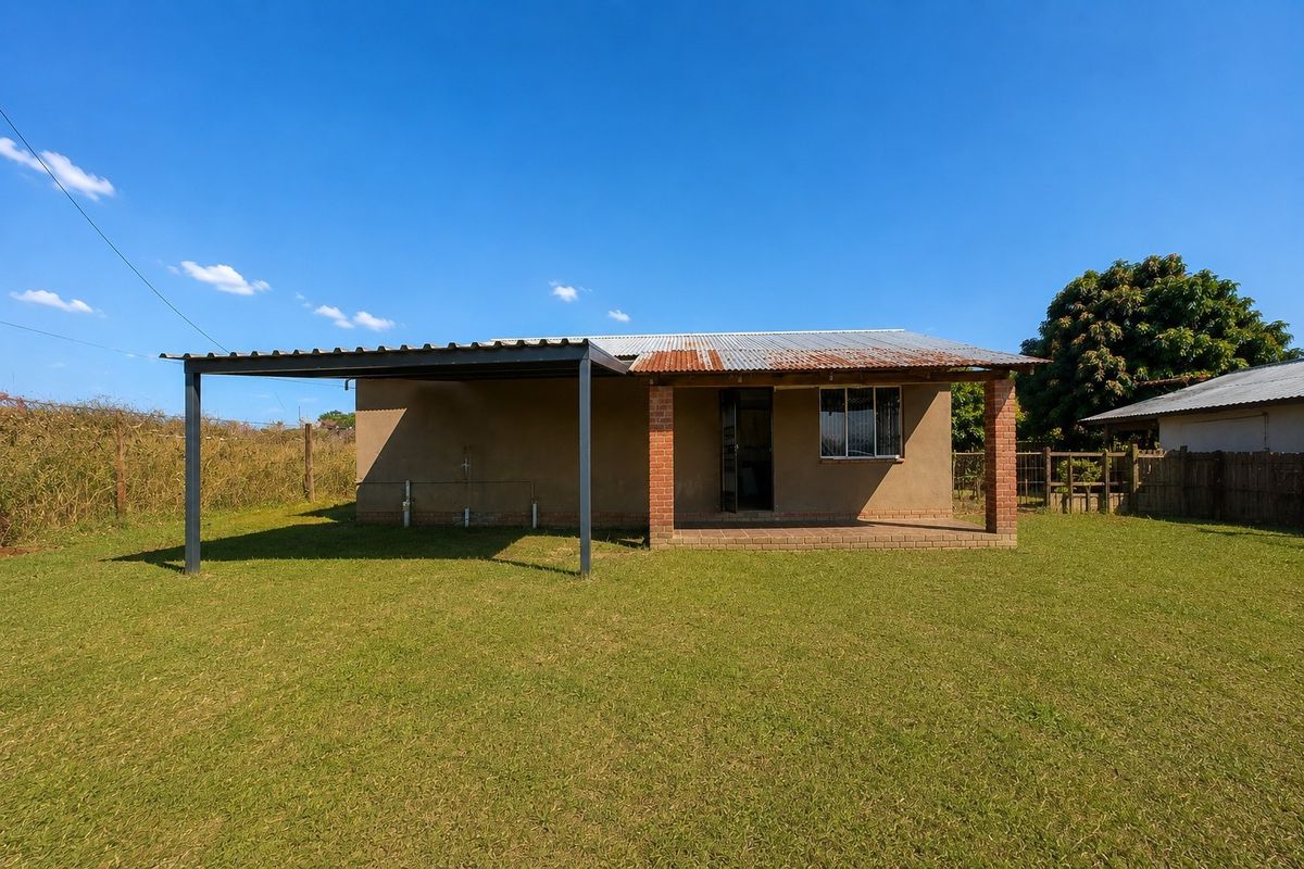 Rear view of a modest house featuring a corrugated metal roof, brick pillars, and an attached carport structure set within a large grassed yard.