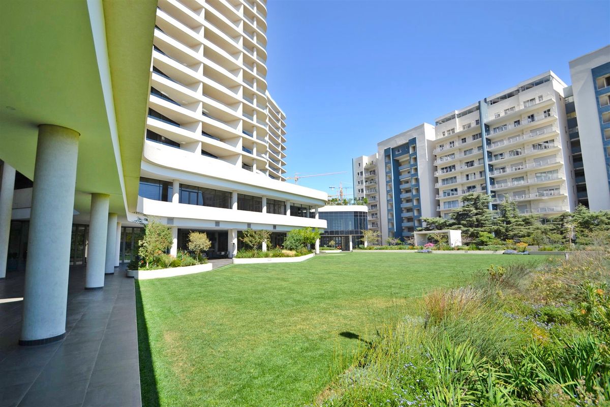 A well-maintained communal garden area featuring a large lawn, landscaped beds, and a covered walkway connecting to the modern apartment building complex.