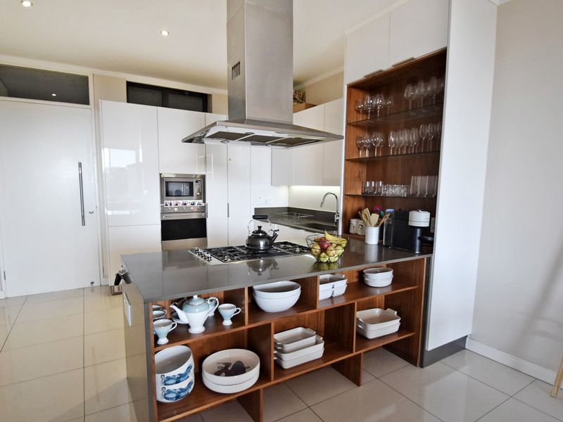 Modern kitchen featuring a central island with gas cooktop, stainless steel extractor, built-in appliances, and sleek white cabinetry with integrated shelving.
