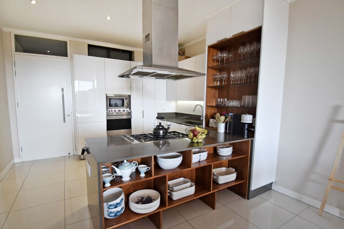 Modern kitchen featuring a central island with gas cooktop, stainless steel extractor, built-in appliances, and sleek white cabinetry with integrated shelving.