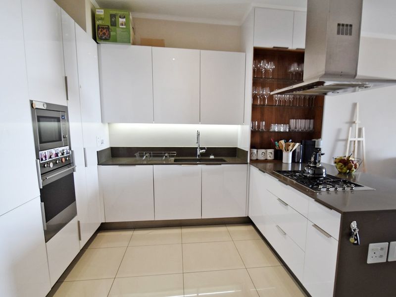 Modern kitchen featuring high-gloss white cabinetry, stone countertops, built-in oven and microwave, gas hob, and integrated shelving with under-cabinet lighting.