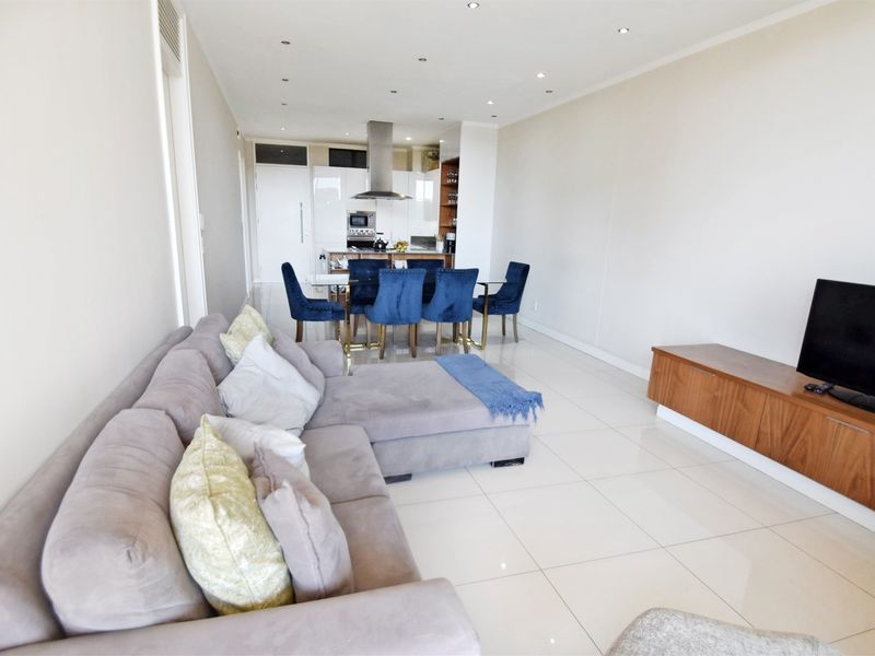 A bright open-plan living and dining area featuring polished porcelain floor tiles, recessed lighting, and a modern kitchen with high-gloss cabinetry and integrated appliances.