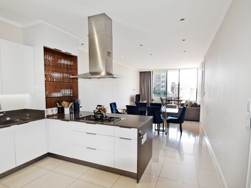 Modern open-plan kitchen and living area featuring sleek white cabinetry, stone countertops, gas hob, stainless steel extractor, and polished tiled flooring leading to a balcony.