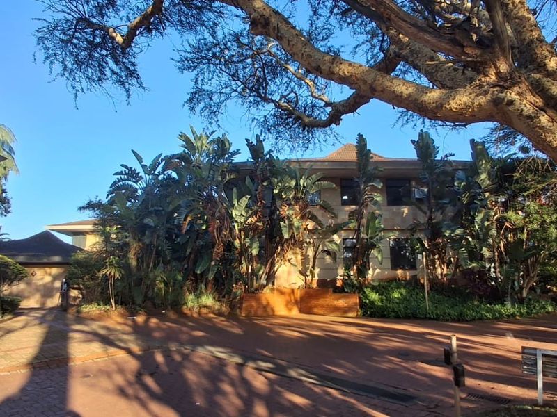 Front view of a multi-story commercial building featuring a paved driveway and established garden landscaping under mature trees.