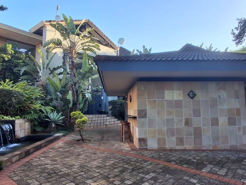 A well-maintained outdoor entertainment area featuring a stone-clad bar structure, paved courtyard, and a tranquil water feature set within a landscaped garden.