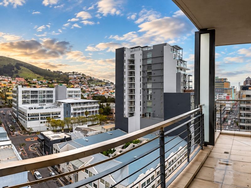 A spacious apartment balcony featuring tiled flooring and a metal railing, offering expansive urban and mountain views.