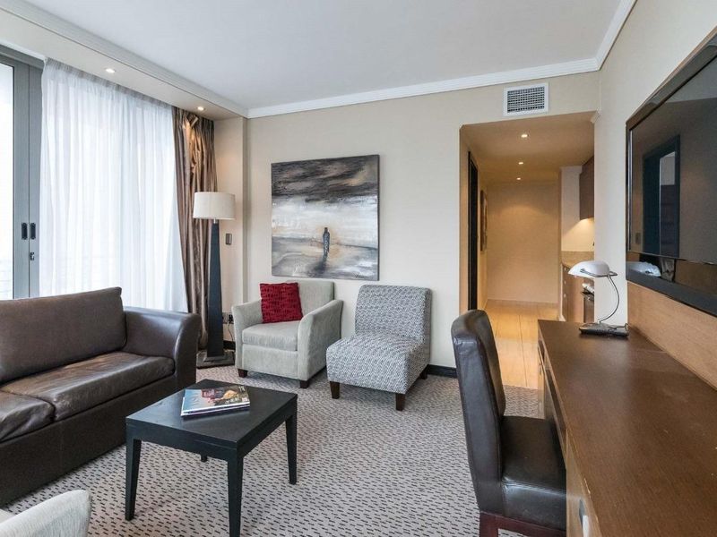 A well-maintained living room featuring carpet flooring, recessed lighting, and a sliding glass door leading to a balcony, with an adjacent kitchen area.