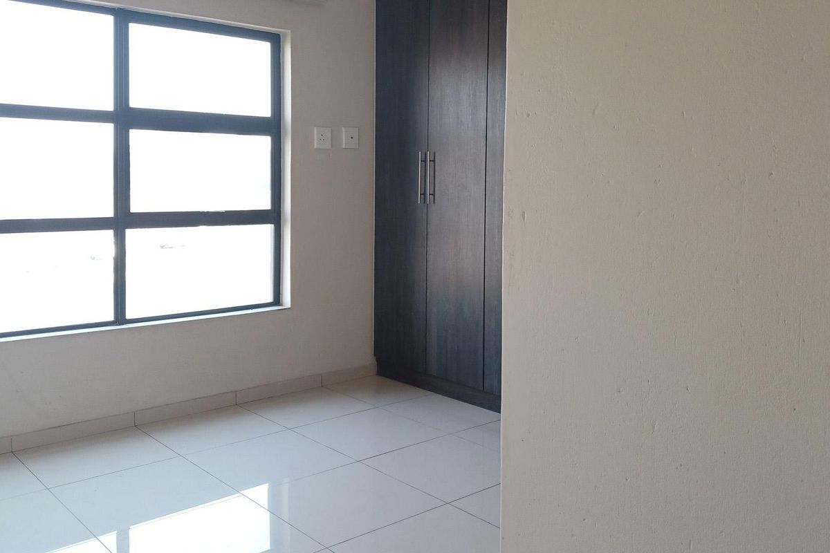 A bright, empty bedroom featuring modern tiled flooring, a large window, and dark wood-finish built-in cupboards.