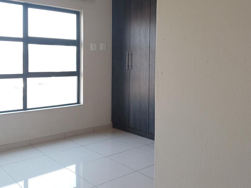 A bright, empty bedroom featuring modern tiled flooring, a large window, and dark wood-finish built-in cupboards.