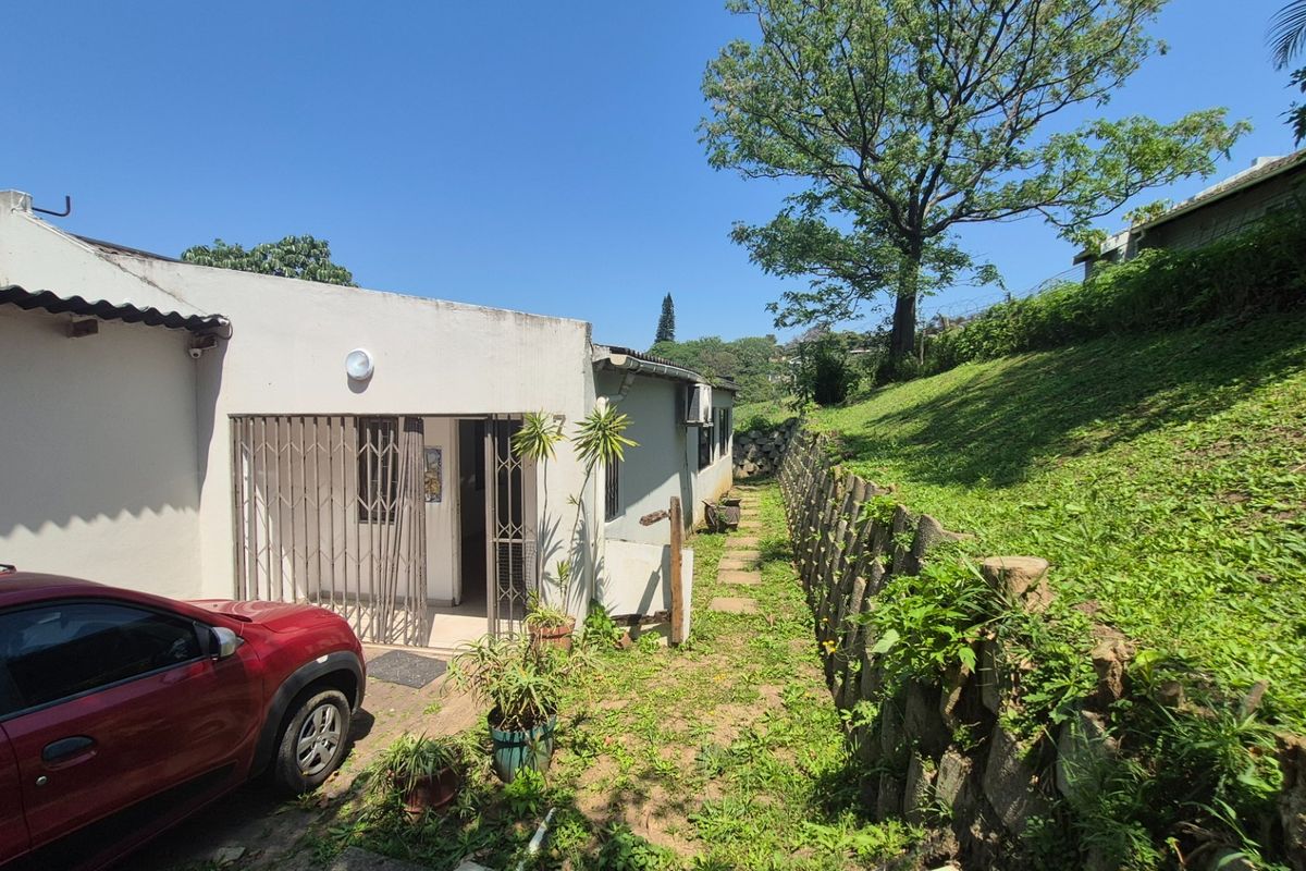 Rear exterior view of the property featuring a paved pathway, security gate, and a stone retaining wall bordering the sloped garden area.