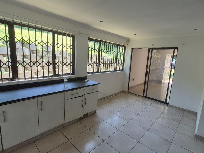 An empty kitchen area featuring tiled flooring, built-in cabinetry with dark countertops, and security bars on the windows, leading to an adjacent room.
