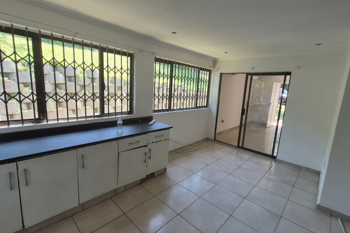 An empty kitchen area featuring tiled flooring, built-in cabinetry with dark countertops, and security bars on the windows, leading to an adjacent room.