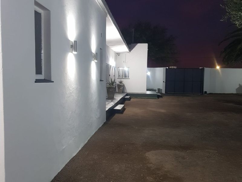 Night view of the property exterior featuring wall-mounted lighting, a secure entrance gate, and a large unpaved driveway area.