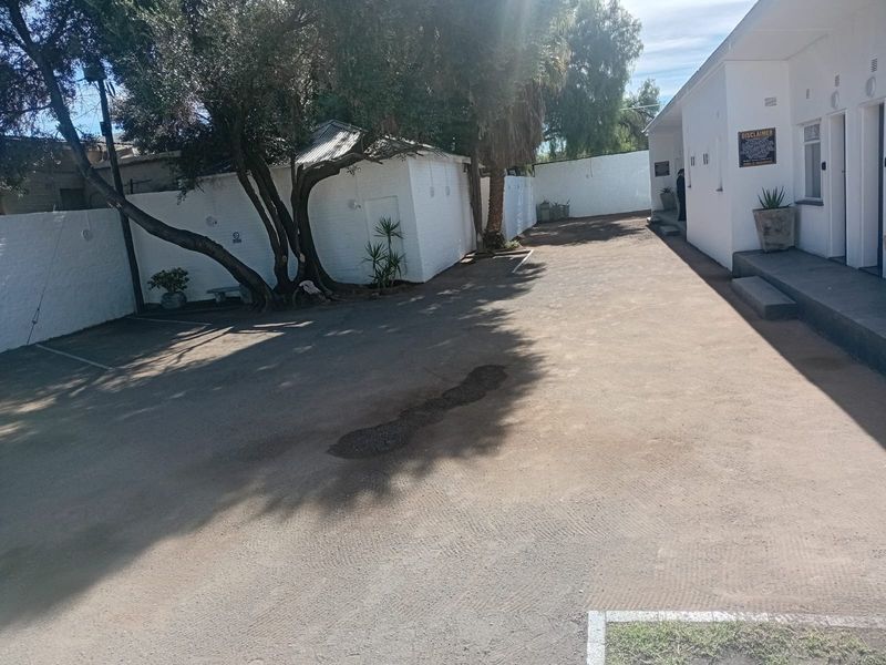 A view of the rear exterior area featuring a paved driveway, boundary walls, and mature trees alongside the guest house building.