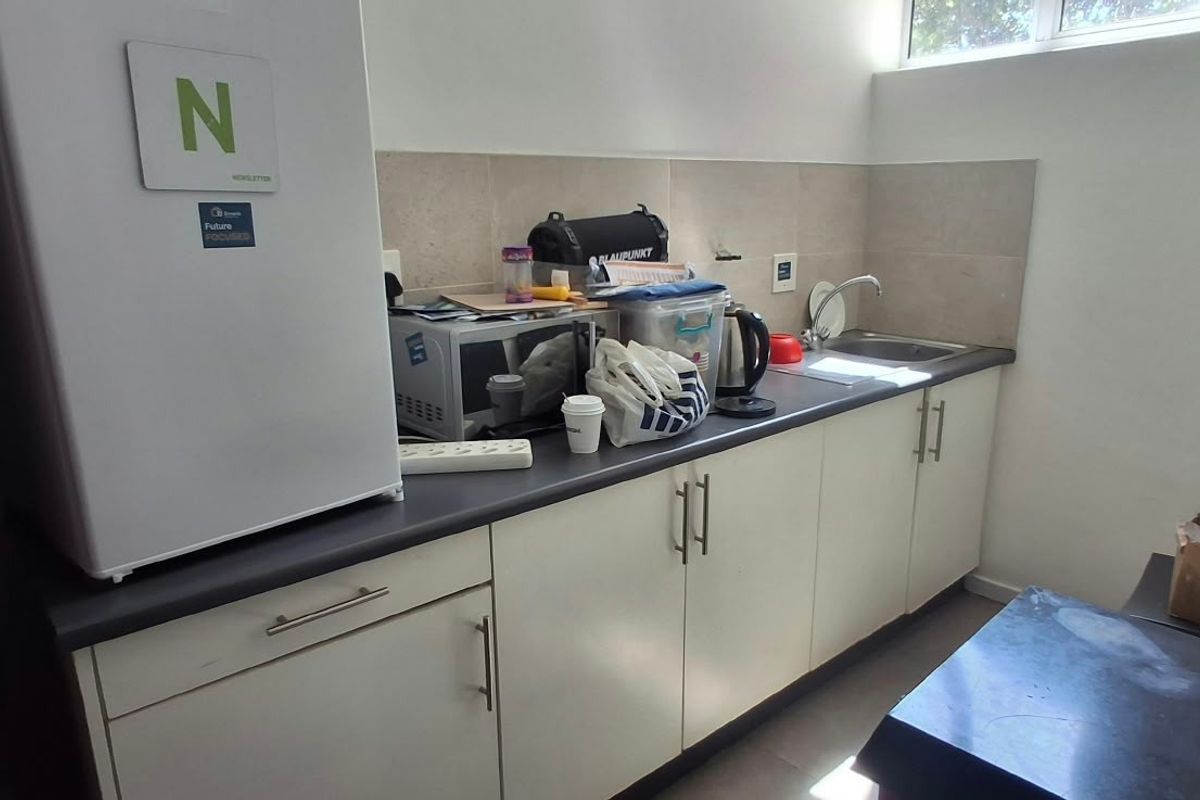 A functional office kitchenette featuring white cabinetry, a dark laminate countertop, stainless steel sink, and tiled backsplash with natural light from a high-level window.
