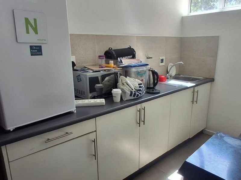 A functional office kitchenette featuring white cabinetry, a dark laminate countertop, stainless steel sink, and tiled backsplash with natural light from a high-level window.