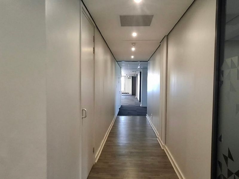 A clean, modern commercial hallway featuring recessed downlighting, wood-look vinyl flooring, and a sleek glass partition wall.